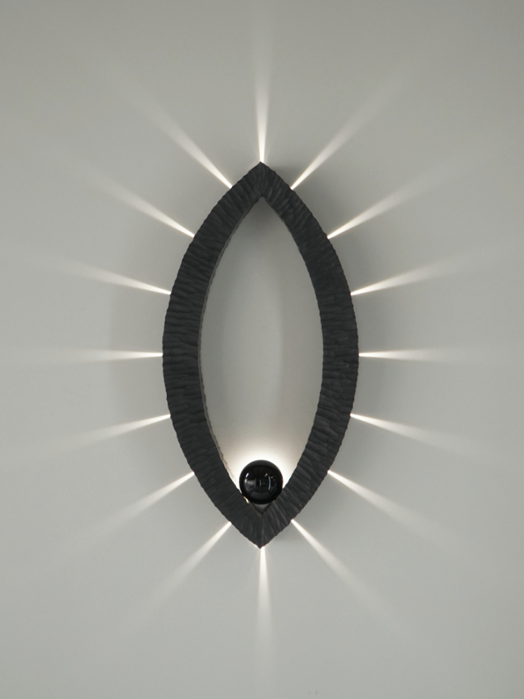 Sculptural wall lamp with an intricate Vesica-inspired geometric design, crafted from wood using fine cabinetmaking techniques. Mounted on a wall, it features a soft, dimmable LED light and a discreet button at the bottom for brightness control.
