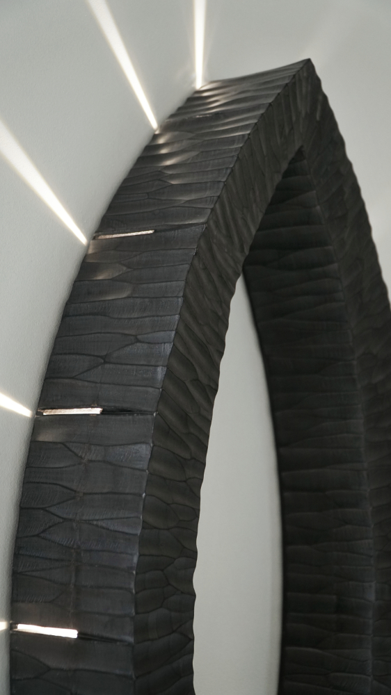 Sculptural wall lamp with an intricate Vesica-inspired geometric design, crafted from wood using fine cabinetmaking techniques. Mounted on a wall, it features a soft, dimmable LED light and a discreet button at the bottom for brightness control.