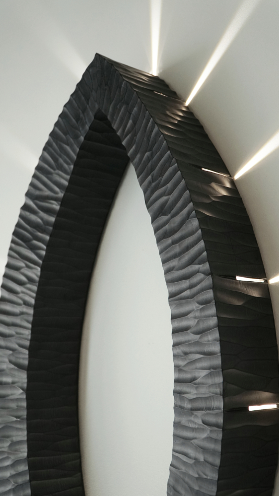 Sculptural wall lamp with an intricate Vesica-inspired geometric design, crafted from wood using fine cabinetmaking techniques. Mounted on a wall, it features a soft, dimmable LED light and a discreet button at the bottom for brightness control.