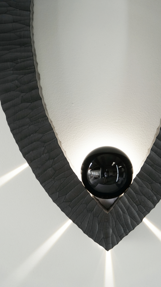 Sculptural wall lamp with an intricate Vesica-inspired geometric design, crafted from wood using fine cabinetmaking techniques. Mounted on a wall, it features a soft, dimmable LED light and a discreet button at the bottom for brightness control.