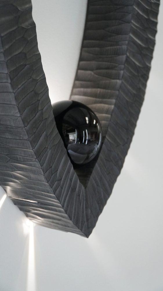 Sculptural wall lamp with an intricate Vesica-inspired geometric design, crafted from wood using fine cabinetmaking techniques. Mounted on a wall, it features a soft, dimmable LED light and a discreet button at the bottom for brightness control.