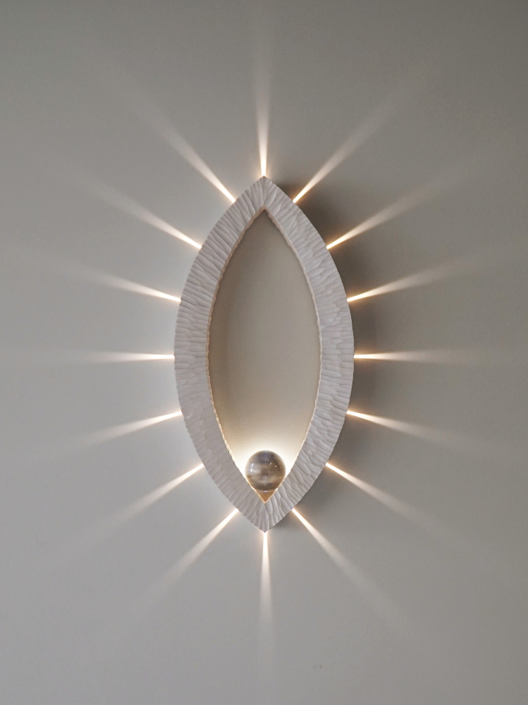 Sculptural wall lamp with an intricate Vesica-inspired geometric design, crafted from wood using fine cabinetmaking techniques. Mounted on a wall, it features a soft, dimmable LED light and a discreet button at the bottom for brightness control.