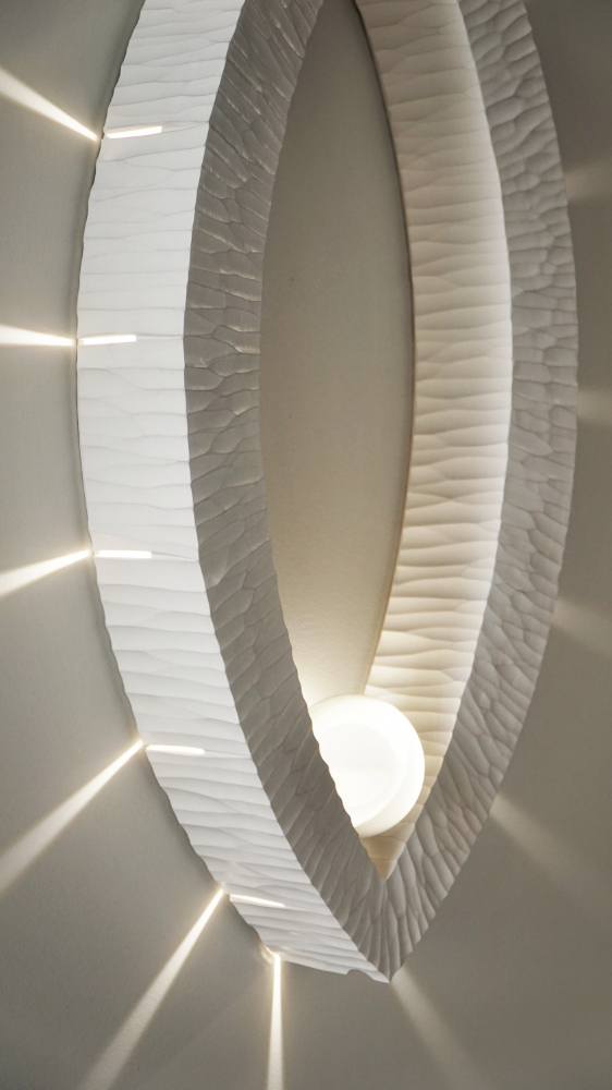 Sculptural wall lamp with an intricate Vesica-inspired geometric design, crafted from wood using fine cabinetmaking techniques. Mounted on a wall, it features a soft, dimmable LED light and a discreet button at the bottom for brightness control.