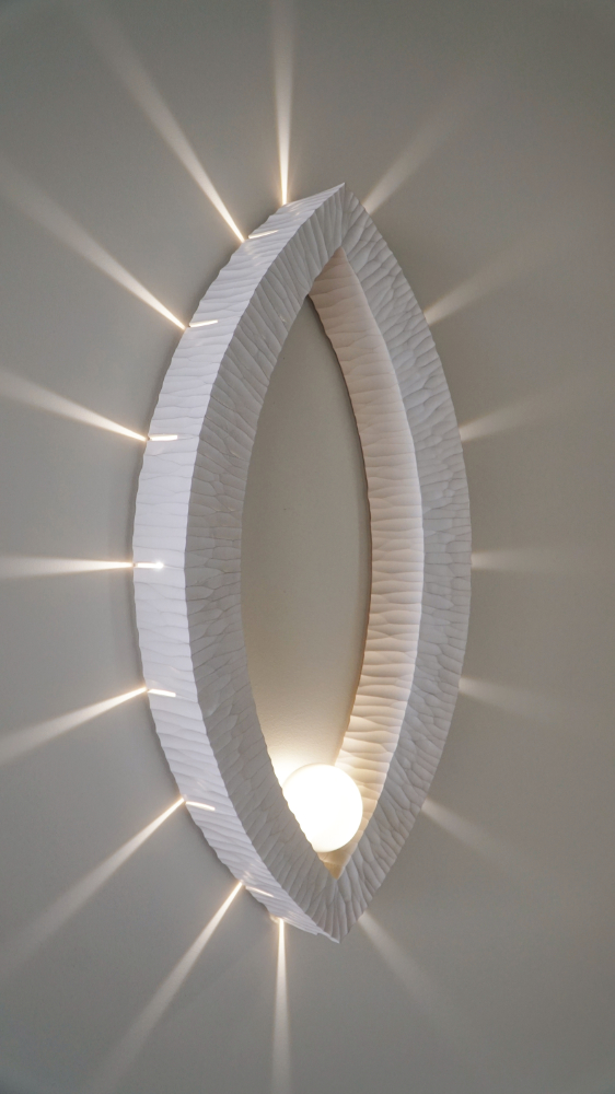 Sculptural wall lamp with an intricate Vesica-inspired geometric design, crafted from wood using fine cabinetmaking techniques. Mounted on a wall, it features a soft, dimmable LED light and a discreet button at the bottom for brightness control.